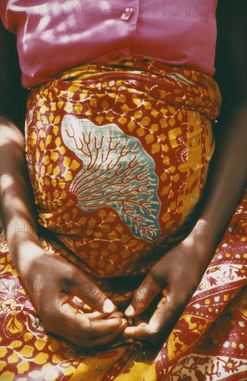 GHANA, Volta Region, Cropped shot of woman thirty six weeks pregnant wearing brightly coloured sarong and with her hands folded in her lap beneath her belly.