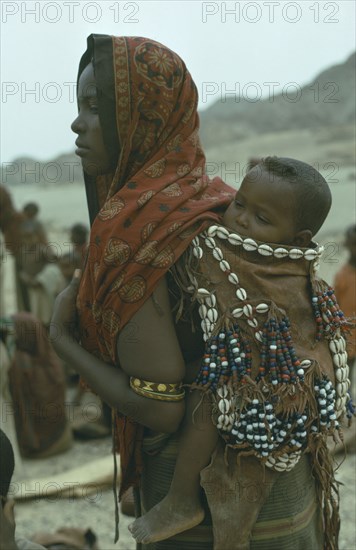 ETHIOPIA, Children, Carrying, Danakil tribeswoman carrying baby on her back in sling decorated with cowrie shells used for barter.