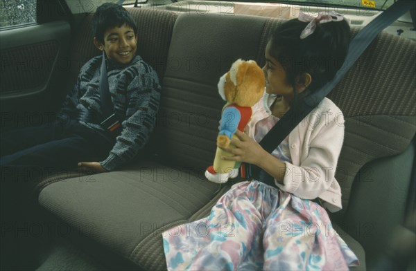 ENGLAND, London, Children, Two Bengali children in back seat of car wearing seat belts.  Young girl showing teddy bear to boy.