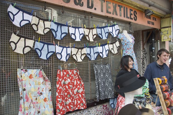 ENGLAND, East Sussex, Brighton, A North Lanes market stall selling hats and clothes with a  large colourful display of underwear behind two young smiling stall holders.