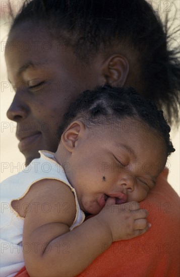 WEST INDIES, St Lucia, Portrait of young mother holding baby girl asleep on her shoulder with thumb in her mouth.