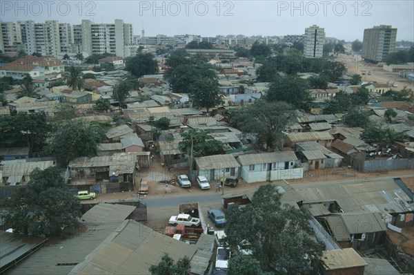 ANGOLA, Luanda, Cars parked next to houses along a road with high rise apartment blocks in the distance.