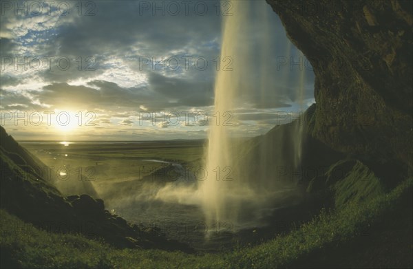 ICELAND, Seljalandfoss, View from behind waterfall in low golden sunlight through drifting cloud.