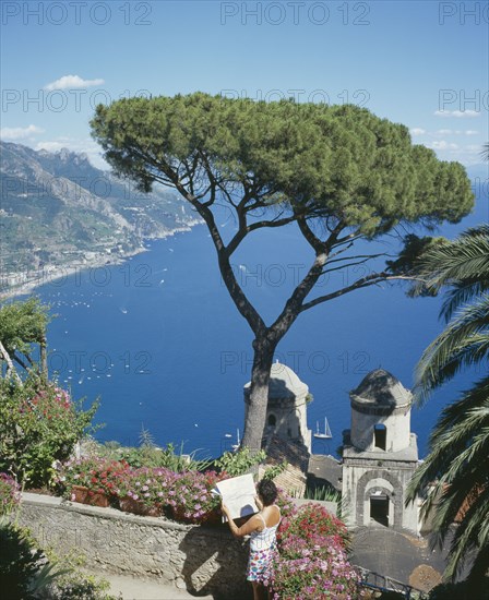 ITALY, Campania, Ravello, "Villa Rufolo, view from gardens to Maiori, Salerno. Tree in the foreground, boats in the bay. Woman reading a map."