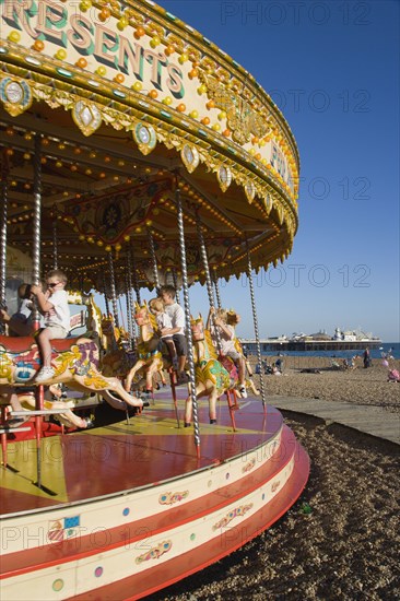 ENGLAND, East Sussex, Brighton, Children riding on a fairground carrousel on the beach with Brighton Pier in the distance