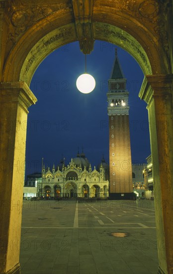 ITALY, Veneto, Venice, Piazza di San Marco.  Part view of square with Basilica di San Marco and the Campanile framed by stone archway with central circular lamp illuminated at night.