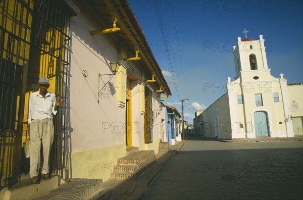 CUBA, Camaguey, Pastel coloured houses lining Playa de San Juan de Dios and Iglesia San Juan de Dios with man opening metal screen of house doorway in the foreground.