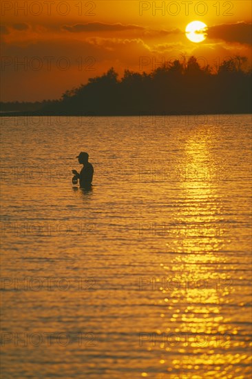 CUBA, Bay of Pigs, Line fisherman standing waist deep in water silhouetted against deep orange sunset reflected in water surface.