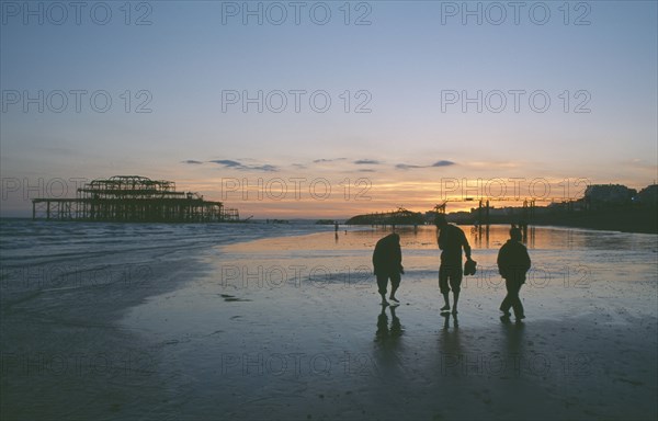 ENGLAND, East Sussex, Brighton, The ruined West Pier at sunset with low tide and a group of people walking together along beach.