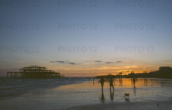 ENGLAND, East Sussex, Brighton, The ruined West Pier at sunset with low tide and a group of people walking a dog together along beach.