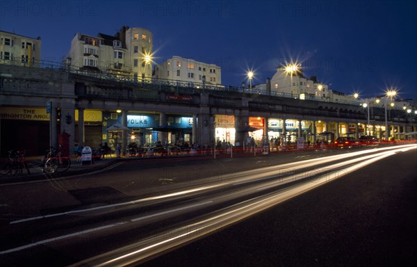 ENGLAND, East Sussex, Brighton, Seafront Promenade shops and bars illuminated at night with traffic in motion blur in the foreground.