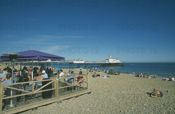ENGLAND, East Sussex, Eastbourne, Beach Cafe with people sat on chairs under parasols and the pier in the distance.
