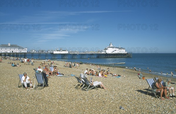 ENGLAND, East Sussex, Eastbourne, View across shingle beach with people sat on deckchairs towards the pier.