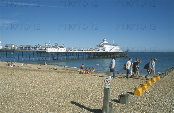 ENGLAND, East Sussex, Eastbourne, View across shingle beach towards pier with a group of people walking past and a No Dogs sign on a post in the foregound.