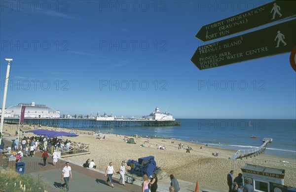 ENGLAND, East Sussex, Eastbourne, View across busy seafront towards shingle beach and pier with direction signs for tourist attractions in the forground.