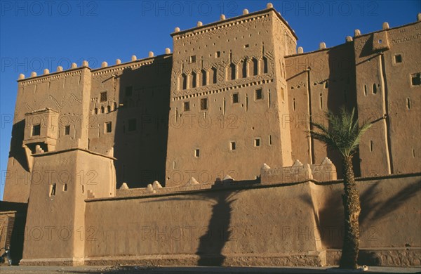 MOROCCO, Ouarzazate, Kasbah Taorirt .  Nineteenth century kasbah of the el-Glaoui dynasty.  Exterior view with shadow from palm tree in foreground.