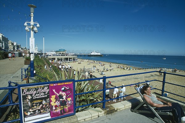 ENGLAND, East Sussex, Eastbourne, View from promenade towards beach and bandstand with a woman sat on deckchair next to blue railings in the forground.