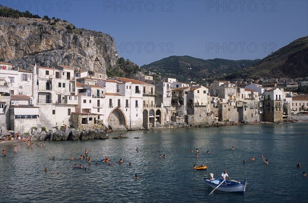 ITALY, Sicily, Palermo, Cefalu. View across people swimming in the sea next to sandy beach with a man rowing a small boat in the foreground overlooked by waterfront apartments and hills behind