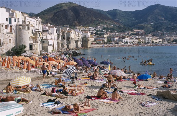 ITALY, Sicily, Palermo, Cefalu. View across busy sandy beach with sunbathers on sand and swimming in the sea overlooked by waterfront apartments and hills