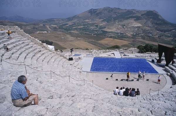 ITALY, Sicily, Segesta, Graeco Roman Amphitheatre. Man sat on steps looking down over stage area with a group of people sat on the steps at the bottom having their photograph taken near stage lighting equipment