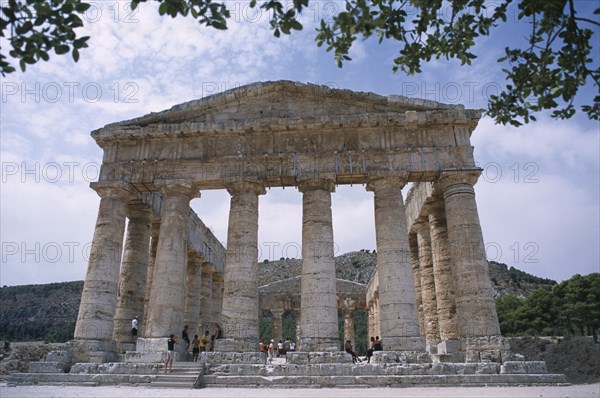 ITALY, Sicily, Segesta, Doric Temple. Ruins of the ancient city. Colonnaded structure framed by tree branches with visitors walking around