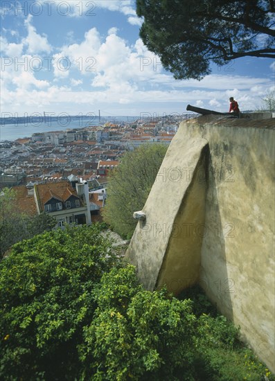 PORTUGAL, Lisbon, Visitor sitting on fortified walls beside cannon of Castelo de Sao Jorge  with rooftop view across city.
