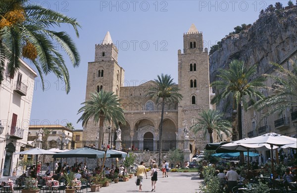 ITALY, Sicily, Palermo, Cefalu. Pathway towards II Duomo Norman Cathedral between cafes with people sat at table and chairs near palm trees