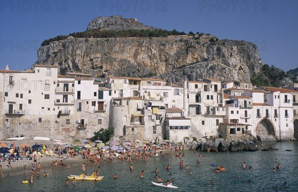 ITALY, Sicily, Palermo, Cefalu. View across the sea with people swimming in the water and sunbathers on busy sandy stretch of beach towards waterfront apartments overlooked by hills behind