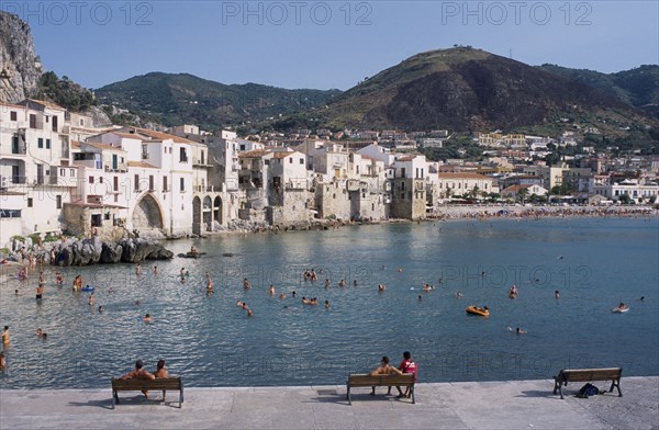 ITALY, Sicily, Palermo, Cefalu. View from stone walls with people sat on benches towards sunbathers swimming in the sea overlooked by waterfront apartments and hills behind