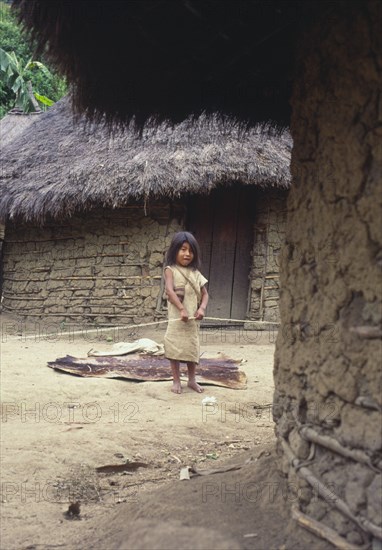 COLOMBIA, Santa Marta, Sierra Nevada, "Young Cogi girl holding cotton yarn, standing in-between two huts."