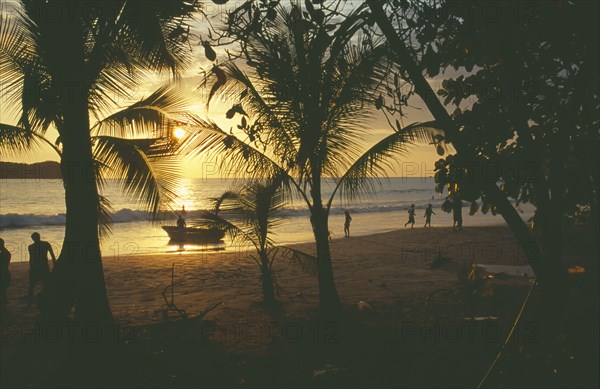COSTA RICA, Puntarenas Province, Manuel Antonio , View from beach at sunset through palm trees towards water with people on sand.