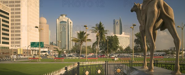 UAE, Dubai, "Camel statue in Al-Ittihad Square, people sat on grass. Palm trees and car park in distance."