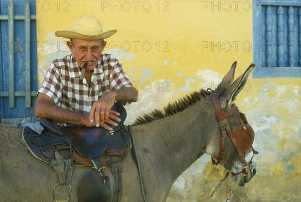 CUBA, Trinidad, Portrait of elderly Cuban man smoking a cigar and leaning on saddle of blinkered donkey.