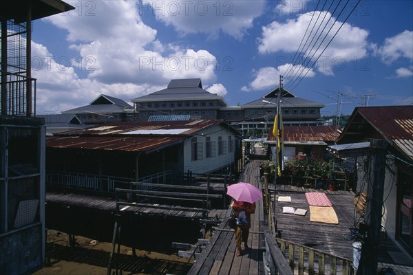 BRUNEI, Bandar Seri Begawan, Kampong Ayer residential district.  Sprawling stilt village on the Brunei River. Person with sun umbrella walking across wooden bridge between houses.