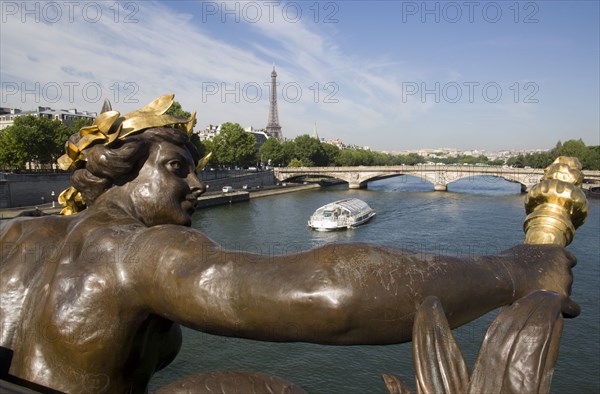 FRANCE, Ile de France, Paris, Bronze statue of a woman holding a flame on the Pont Alexandre III bridge over the River Seine with a Bateaux mouches pleasure boat with tourists passing below and the Eiffel Tower in the distance