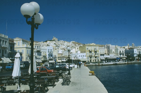GREECE, Cyclades Islands, Syros, Ermoupolis. The town with benches and cafes at the waters edge with the orthodox church “Anastasi” on top of the hill bedind.