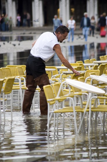 ITALY, Veneto, Venice, Aqua Alta High Water flooding in St Marks Square with waiter wearing waders and preparing restaurant tables