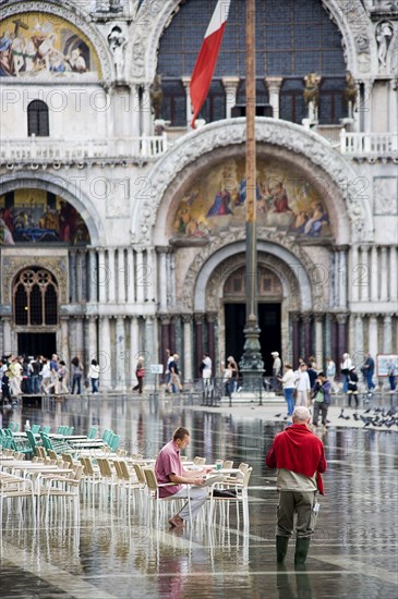 ITALY, Veneto, Venice, Aqua Alta High Water flooding in St Marks Square showing St Marks Basilica at the end of the flooded piazza with an artist painting a watercolour seated at a table in the water