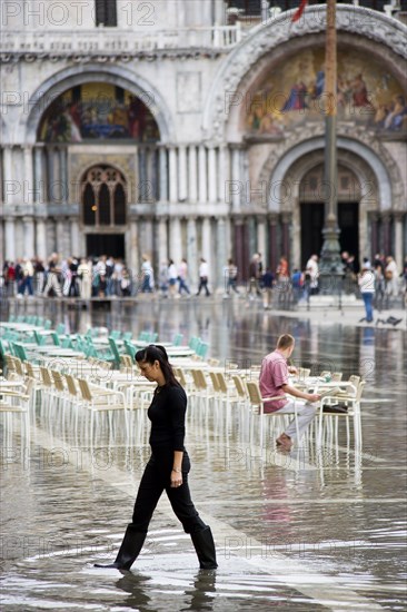 ITALY, Veneto, Venice, Aqua Alta High Water flooding in St Marks Square showing St Marks Basilica at the end of the flooded piazza with an artist painting a watercolour seated at a table in the water and local female resident wearing wellington boots walking in the water