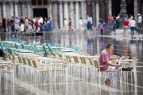 ITALY, Veneto, Venice, Aqua Alta High Water flooding in St Marks Square showing St Marks Basilica at the end of the flooded piazza with an artist painting a watercolour seated at a table in the water