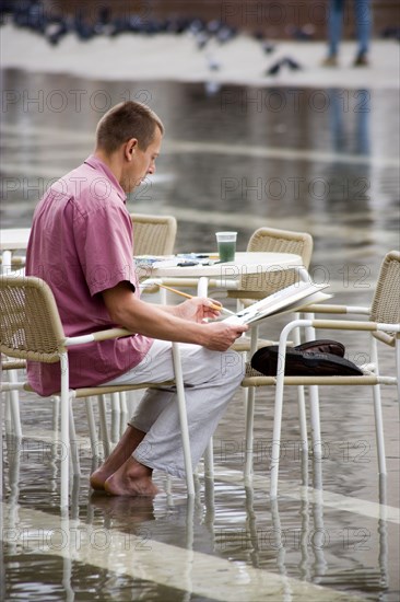 ITALY, Veneto, Venice, Aqua Alta High Water flooding in St Marks Square with an artist painting a watercolour seated at a table in the water Paul Seheult