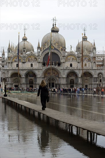 ITALY, Veneto, Venice, Aqua Alta High Water flooding in St Marks Square showing St Marks Basilica at the end of the flooded piazza and a resident walking to work on an elevated walkway