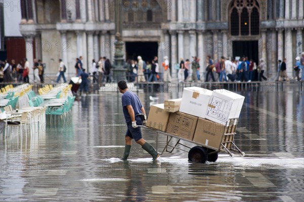 ITALY, Veneto, Venice, Aqua Alta High Water flooding in St Marks Square with a delivery man pulling a trolley of goods through the water with St Marks Basilica at the end of the flooded piazza