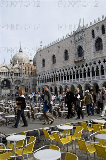 ITALY, Veneto, Venice, Aqua Alta High Water flooding in St Marks Square with tourists walking on an elevated walkway through restaurant tables by the Doges Palace with St Marks Basilica beyond