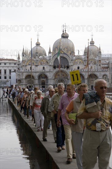 ITALY, Veneto, Venice, Aqua Alta High Water flooding in St Marks Square A party of tourists walking on an elevated walkway above the water with St Marks Basilica at the far end of the piazza