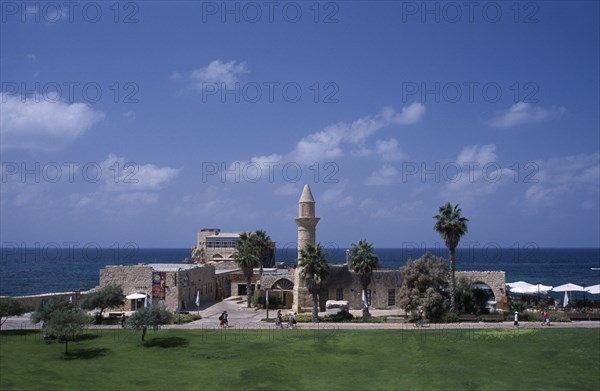 ISRAEL, Caesarea, The Herodian Harbour with eastern view of stone buildings with palm trees and visitors walking past along path.