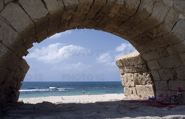 ISRAEL, Caesarea, Roman Aqueduct with view through a single arch towards sunbathers on the beach next to the Mediterranean Sea