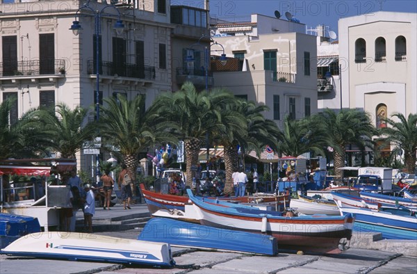 ITALY, Sicily, Palermo, Mondello. Fishing boats moored next to the promenade overlooked by buildings with people walking along path near palm trees