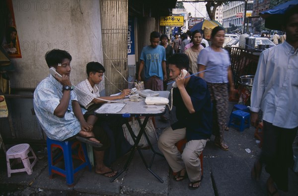 MYANMAR, Yangon, Public telephone exchange.  Young men using telephones set up on folding table on busy street.