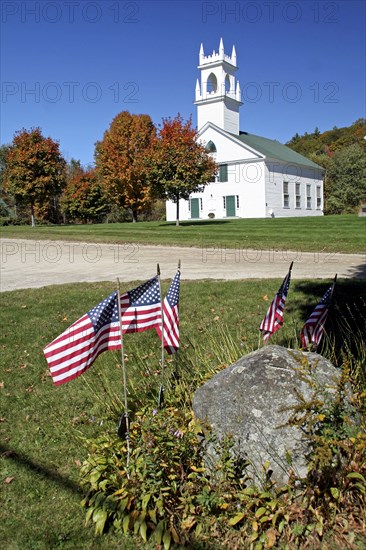 USA, New Hampshire, Autumn foliage, White wooden church and autumnal trees with American Stars & Stripes flags in the foreground.
